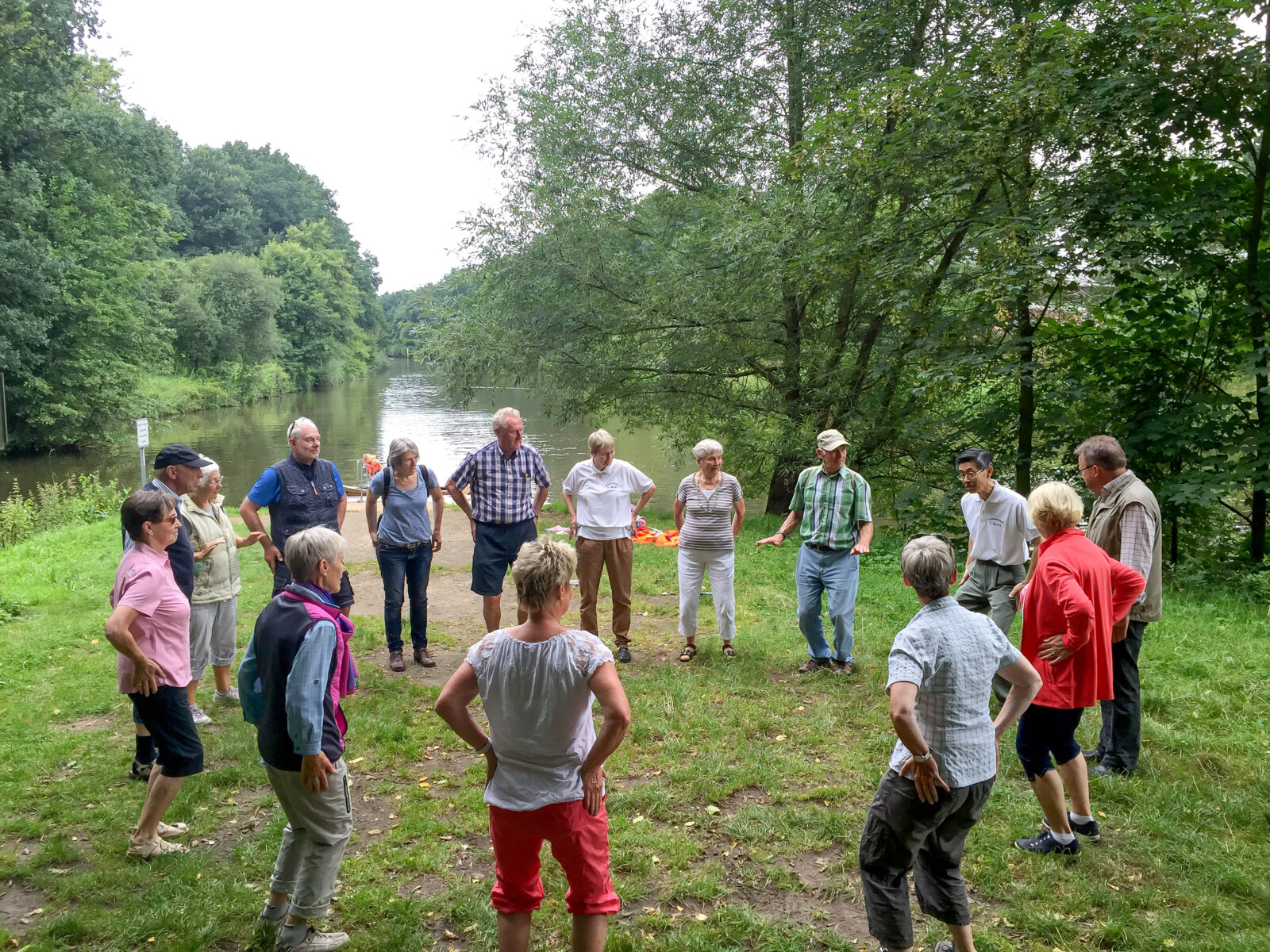 An einem Gewässer stehen Menschen in einem Kreis und hören dem Gruppenleiter zu
