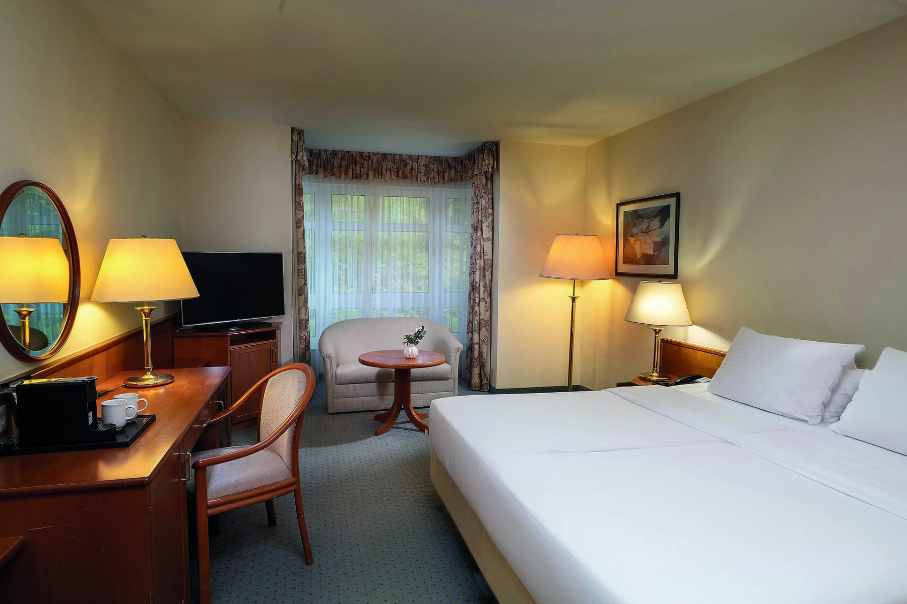 Hotelzimmer mit Doppelbett, Schreibtisch, Flachbildschirm und kleinem Sofa vor bodentiefen Fenstern mit Gardinen.