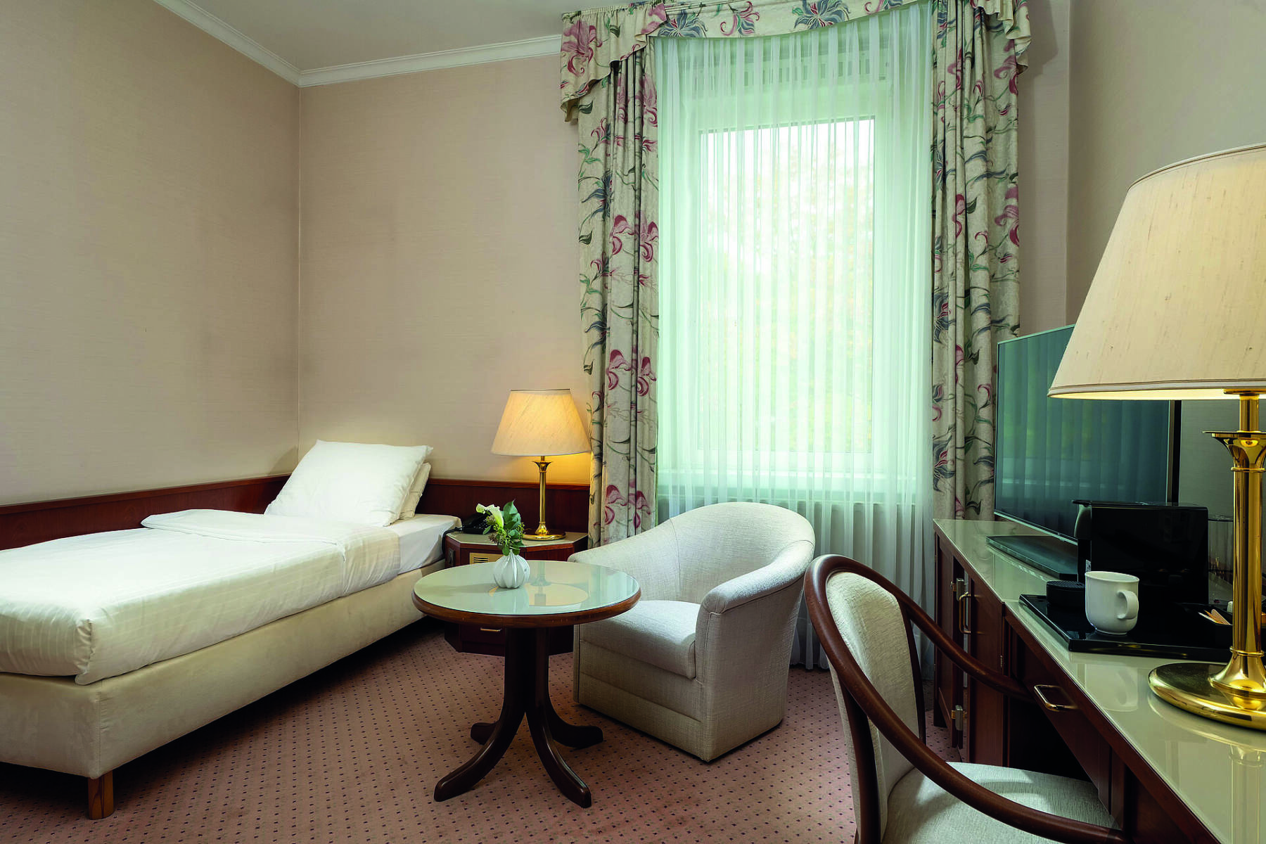 Hotelzimmer mit Einzelbett, Sessel, kleinem runden Tisch, Schreibtisch mit Stuhl, Fernseher und zwei Tischlampen.