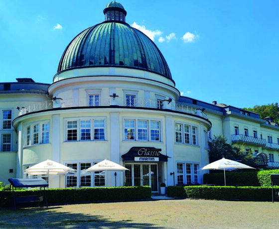 Rundes Gebäude mit kupfergrünem Kuppeldach und weißer Fassade vor blauem Himmel mit Sonnenschirmen und Bänken im Vordergrund