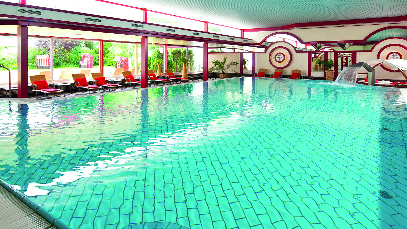 Schwimmbecken mit türkisfarbenem Wasser. Große Fenster mit Blick ins Grüne. Um das Becken herum stehen Liegestühle.
